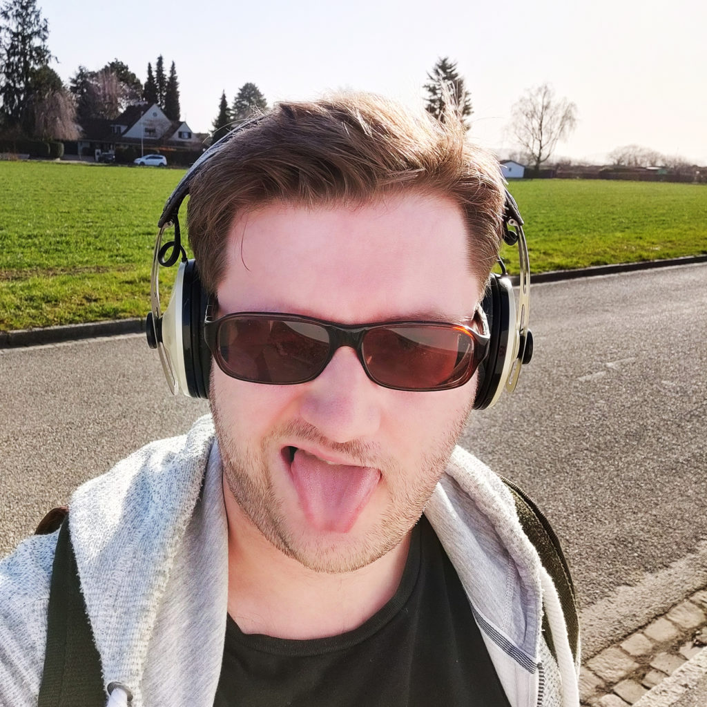 Ein Selbstporträt. Ich trage Sonnenbrille und Kopfhörer, stehe auf einer Straße vor einer sonnenbeschienenen Wiese, und strecke dem Betrachter versucht lässig die Zunge heraus.